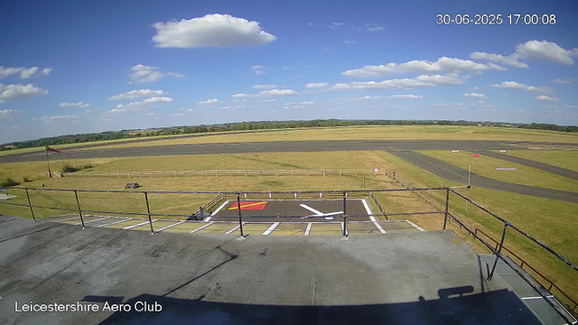 A wide view of an airfield runway during daytime with a clear blue sky and scattered clouds. In the foreground, there is a small parking area with designated parking slots and a red-and-white striped section. A flagpole with a flag is visible near the parking area, and a bench is placed on the grass. The horizon shows a flat landscape with distant trees and fields. The timestamp reads "30-06-2025 17:00:08" and the location is marked as "Leicestershire Aero Club."