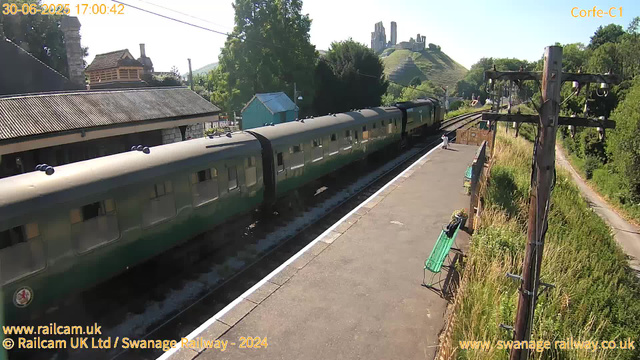 A green passenger train is stopped at a small rural station platform. The platform has a bench and a person sitting on it. To the right, there are wooden utility poles with electrical wires extending along the train tracks. In the background, there are trees, a small building, and a hill with the ruins of an old castle or fortress on top. The sky is clear and sunny. The timestamp on the image shows the date as June 30, 2025, at 17:00:42.