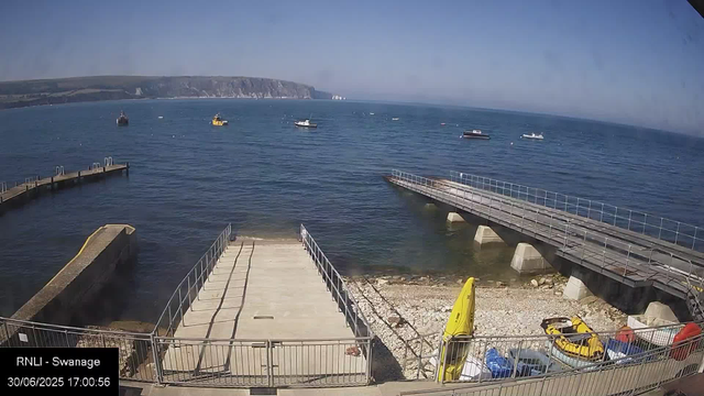 A view of a seaside dock area with a concrete pier extending into the water. Several boats are anchored in the distance on a calm, blue sea. To the right, there's a ramp leading into the water, supported by concrete supports. The shoreline below consists of small rocks and pebbles, with some yellow safety equipment and a few small vehicles parked near the fence in the foreground. In the background, there are white chalk cliffs along the coastline under a clear, sunny sky.