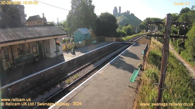 A small railway station platform with a building on the left, a green bench, and a small greenhouse nearby. The railway tracks run through the scene, curving to the right, with greenery and trees lining the area. In the background, there is a hill with the ruins of a castle or historic structure on top. The sky is clear and sunny. There are utility poles with wires running along the right side of the scene. The timestamp shows the date as June 30, 2025, at 6:00 PM.