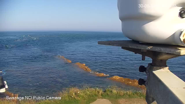 A view of the ocean with a rocky outcrop extending into the water, seen from a webcam mounted on a platform. The water is calm with gentle waves, and the sky is clear and blue. The corner of a white lighthouse or similar structure is visible on the right side of the image. The image has a timestamp in the top right corner and a label at the bottom left that reads "Swanage NCI Public Camera."