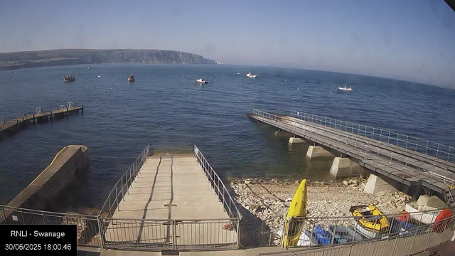 A view of a harbor with several small boats floating on calm water. There is a concrete pier extending into the water, with a ramp leading down to the shore. On the rocky beach area, there are yellow life jackets, a kayak, and some parked cars. In the background, cliffs line the horizon under a clear blue sky.