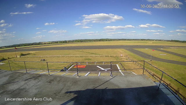 A clear daytime view from a webcam at Leicesterhire Aero Club, showing an open tarmac area with a handicapped parking spot in the foreground, a grassy field, and a runway extending across the middle ground. The sky is mostly clear with some scattered clouds. A wind sock is visible on the left side, and the horizon reveals distant trees and fields. The timestamp in the upper right corner indicates the date as June 30, 2025, and the time as 18:00:50.