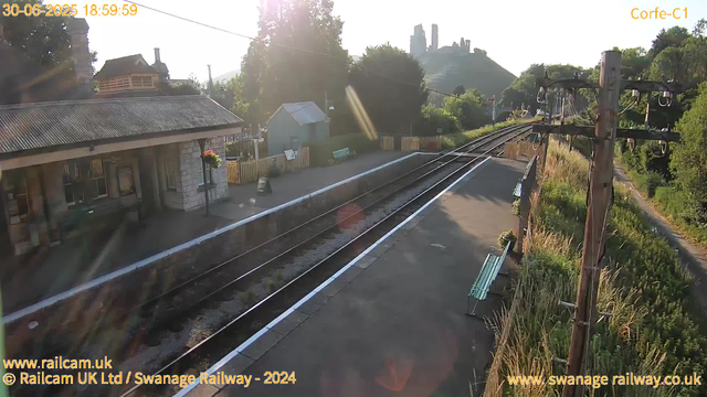 A small railway station platform with a building on the left, flowers hanging from the wall, and a sign that says "WAY OUT." There are railroad tracks running through the center, with a set of overhead electrical wires and poles on the right. In the background, there's a small green building, trees, and a hillside with ruins or structures at the top, all illuminated by the late afternoon or early evening sunlight.
