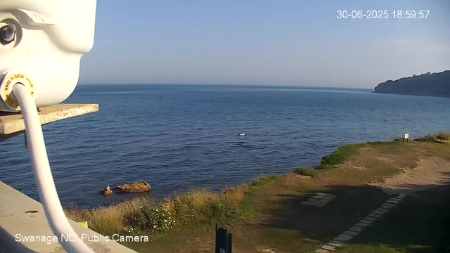 A view of the ocean coastline on a clear day with calm water, a small boat in the distance, a rocky shoreline, and a distant headland on the right. There is a parking area with painted lines in the foreground, and part of a white security camera in the top left corner. The timestamp indicates the date and time as June 30, 2025, 6:59:57 PM.