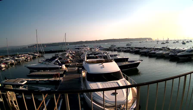 A marina filled with numerous boats and yachts docked along wooden piers, with a calm water surface reflecting the vessels. In the background, there is a distant shoreline under a mostly clear sky.
