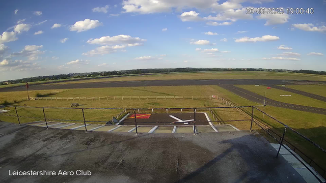 A view from a webcam at Leicestershire Aero Club shows a clear day with blue skies, scattered white clouds, and an expansive grassy field surrounding the runway. The runway is visible in the distance, with a small section of an apron or taxiway in the foreground, including a marked helicopter landing pad. The scene is bright and peaceful, with no visible aircraft or people present. The date and time are displayed in the upper right corner as June 30, 2025, at 7:00 PM.
