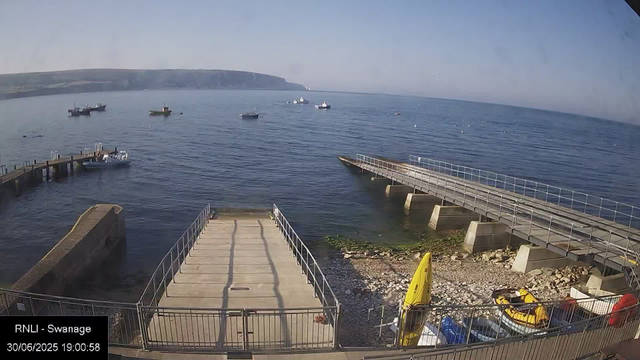 A view of a harbor with several boats floating on calm water. There are two piers extending into the water, one on the left and another on the right. At the bottom of the image, there are boats and equipment, including yellow kayaks and a small red vehicle. The background features a distant coastline with cliffs or hills under a clear sky. The timestamp and location label indicate it was taken on June 30, 2025, at 19:00:58, from RNLI in Swansea.