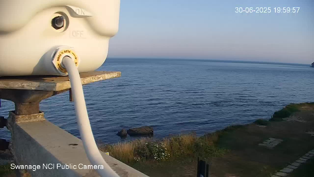 A view from a webcam showing a calm sea with gentle ripples, a clear sky, and distant land in the background. In the foreground on the left, there is a white or light-colored piece of equipment, possibly a camera or sensor, mounted on a weathered wooden platform. The equipment has a yellow dial and a white cable connected to it. The timestamp in the top right corner reads "30-06-2025 19:59:57," and text in the bottom left indicates the location as "Swanage NCI Public Camera."