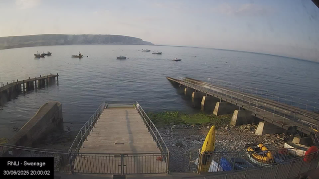 A tranquil coastal scene with a small pier extending into calm water. Several boats are anchored in the distance, and a larger boat moves across the water. On the right side, a floating dock or barrier is visible. The shoreline below has some rocks and green algae, with a yellow boat and other small vehicles parked nearby. The sky is clear with a slight haze near the horizon, suggesting early evening. In the bottom left corner, there is a timestamp indicating the date and time as 30/06/2025, 20:00:02.