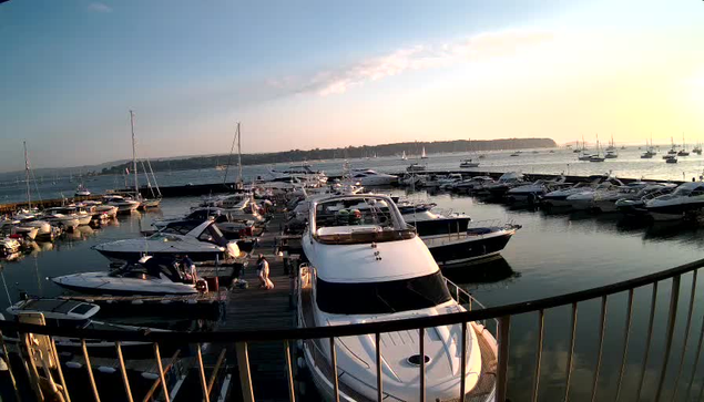 A marina filled with numerous docked boats and yachts, with calm water reflecting the vessels. The sky is partly cloudy with the sun setting, creating a warm glow on the scene. A railing in the foreground partially frames the view.