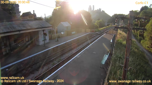 A small railway station platform at sunset, with train tracks running parallel. On the left, there is a building with a corrugated metal roof and a hanging flower basket. On the right, there is a wooden utility pole with electrical wires. Benches are placed along the platform, and a pathway runs alongside the tracks. In the background, a hillside with an ancient castle or ruins is visible, silhouetted against the setting sun. The overall scene is bathed in warm, golden light.