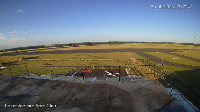 A wide view of an airfield with a small parking area in the foreground, featuring marked spots and a ramp with an arrow. A fence runs along the perimeter of the parking area. Beyond that, there is a grassy area with a windsock on a pole, and a small bench. The main runway stretches across the middle of the image, with a few ground markings, surrounded by green and brown patches of grass. In the distance, there are trees and a mostly clear blue sky with a few scattered clouds. The timestamp shows the date and time as 30-06-2025 at 20:00:47. The bottom left corner has the label "Leicestershire Aero Club."