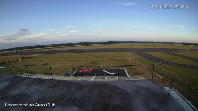 A view from a webcam at Leicesterhire Aero Club shows an open tarmac and grassy airfield with runways in the distance under a clear blue sky with a few scattered clouds. The image is timestamped June 30, 2025, at 8:59 PM. There is a small runway marking area with painted lines and signs in the foreground, and a grassy area with a bench and a pole on the left side. The horizon extends far into the distance with trees and fields.