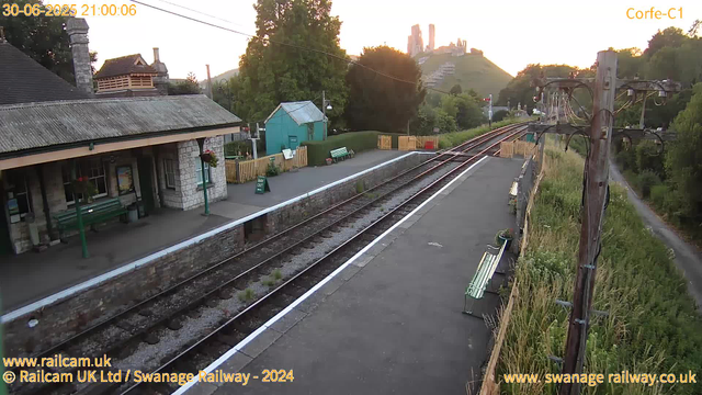 A view of a railway station platform at sunset, with a small building on the left, green benches, a fenced area with a small turquoise shed, and train tracks running through the center. There are power lines and a wooden utility pole on the right, with a grassy area and trees beyond the tracks. In the background, a hill with a castle or fortress ruins is visible on top. The scene appears calm and quiet.