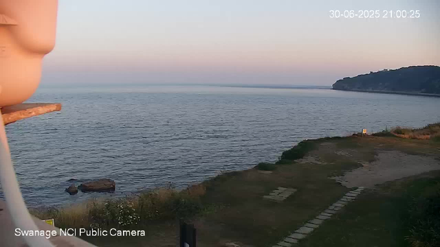 A view of the coastline at dusk, with calm ocean waters stretching towards the horizon. On the right, there is a cliff covered with trees. In the foreground, a grassy area with a pathway leading towards the water, and a small white structure near the shoreline. The sky is clear with the last light of sunset, and the scene appears peaceful and quiet. The timestamp indicates the date as June 30, 2025, at 9:00 PM.