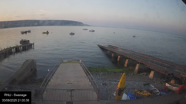 A view of a calm harbor at sunset with several boats floating on the water. There are two piers extending into the water, one on the left and one on the right. The shoreline has some rocky and mossy areas. In the foreground, there is a small concrete slipway with a seagull perched on the railing, and some yellow and blue equipment, including a small boat and a yellow kayak, stored near the railings. The horizon shows distant landforms with a gentle, pastel-colored sky. The timestamp indicates the photo was taken on June 30, 2025, at 21:00:36.