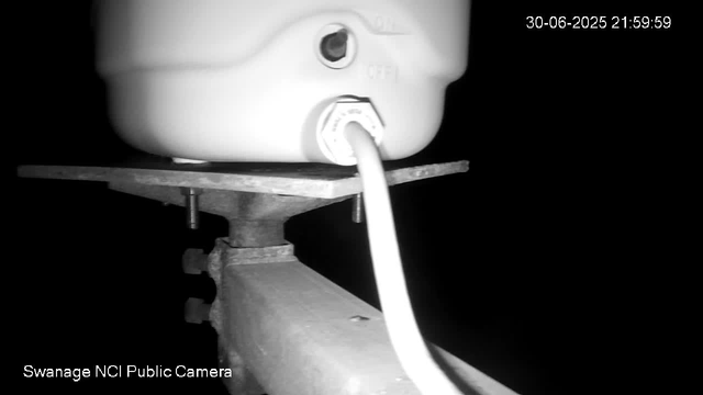 A black-and-white image from a webcam shows a close-up of a white plastic device mounted on a metal pole with bolts. The device has a circular component and a wire connected to it. In the top right corner, the date and time "30-06-2025 21:59:59" are displayed. The bottom left corner contains the text "Swanage NCI Public Camera." The background is dark, indicating it is nighttime.