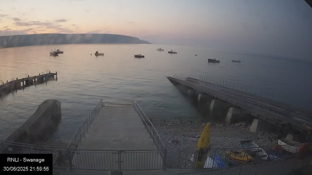 A view of a calm seaside scene at dusk with several boats floating on the water. There is a small pier in the foreground extending into the water, and a larger jetty structure on the right side. The sky has soft pastel colors from the setting sun, and distant landforms are visible on the horizon. At the bottom left, there is a timestamp and location information.