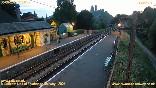 A small railway station platform at dusk with a historic stone station building illuminated by warm lighting. The platform has a few benches, hanging flower baskets, and a green "Way Out" sign. Railway tracks run alongside the platform, curving towards a bridge in the background. In the distance, a hilltop with ruins or old structures is visible against the twilight sky. A wooden utility pole with electrical wires is on the right side of the image, and a footpath runs parallel to the railway on the far right. The scene is calm and quiet, with minimal activity.