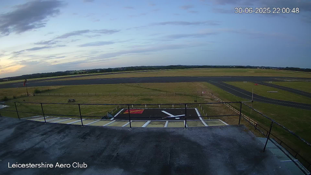 A view from a webcam showing the airfield at Leicestershire Aero Club during early evening. The scene includes the runway extending into the distance, a grassy area with a small flagpole and a flag, a fenced boundary, and a helipad marked with a red, white, and black "H" symbol in the foreground. The sky is partly cloudy with shades of blue and gray, and the timestamp indicates it is 10:00 PM on June 30, 2025.