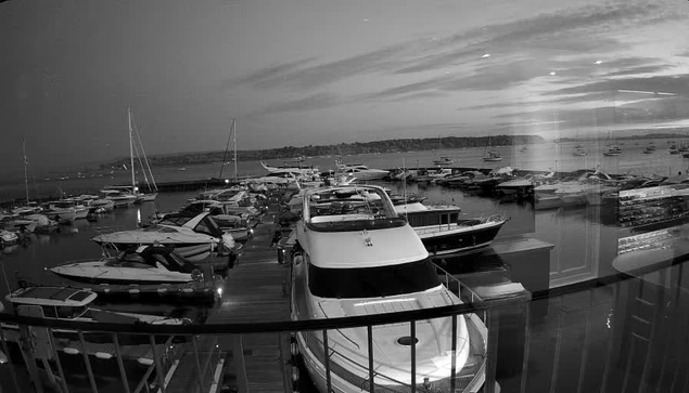 A black-and-white image of a marina with numerous boats docked along the piers. The water is calm, and there are sailboats and motorboats of varying sizes. In the background, there is a view of the distant shoreline and a sky with some scattered clouds.