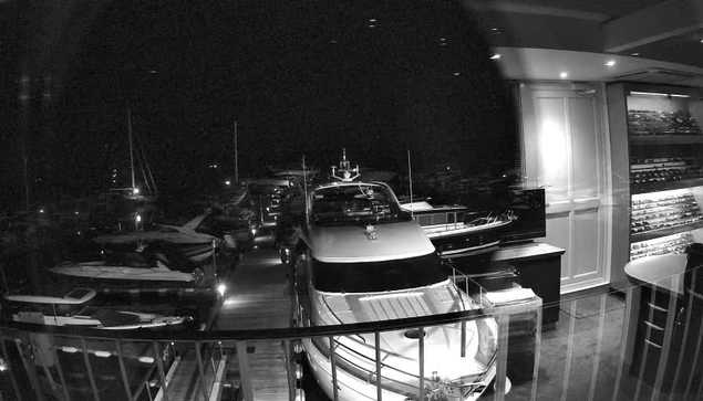 A black-and-white photo shows the interior of a yacht or boat showroom at night. Through a large glass window, several yachts are docked outside on the water, with masts and rigging visible. Inside, there is a display area with a modern yacht, a small cabinet, a counter with some objects on it, and a shelving unit filled with bottles or other items. The room has a curved ceiling with recessed lighting.