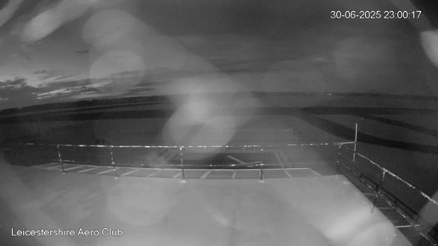 A black and white image taken from a webcam at Leicestershire Aero Club shows an empty parking area with several marked parking spaces and a bicycle rack in the foreground. In the background, there are open fields and a cloudy sky, with some scattered clouds visible. The image appears to be taken at night or in low-light conditions, and there are some light reflections or noise spots in the image. The timestamp in the upper right corner reads "30-06-2025 23:00:17."