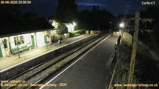 A small train station platform at night, illuminated by a few lights. There are green benches and flower pots with hanging plants along the building. A small green structure, possibly a shed, is on the platform. The station appears quiet with no passengers. Tracks run parallel to the platform, and there is a sign indicating "WAY OUT." The sky is dark, and surrounding area appears to be rural with trees and power lines visible.