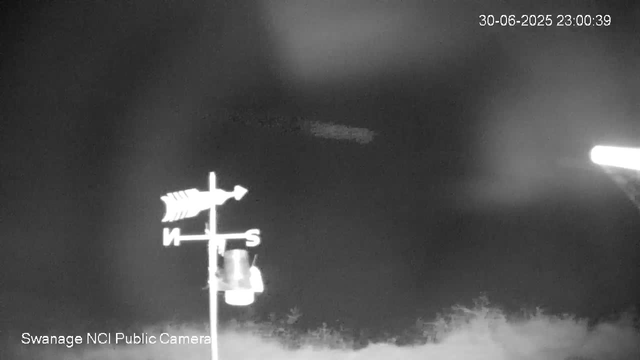 A black-and-white view from a webcam at night shows a weather vane with a fish-shaped arrow pointing to the right. The weather vane includes cardinal directions: North (N), South (S), and an arrow pointing East. The scene is dark with no visible stars or moon, and there is a faint silhouette of trees at the bottom. The timestamp in the upper right corner reads "30-06-2025 23:00:39," and text at the bottom left indicates it is from the "Swanage NCI Public Camera."