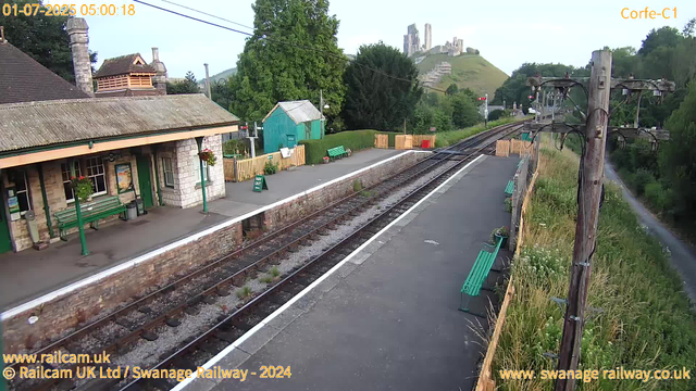 A small train station platform with a station building on the left, featuring a green door, windows, and hanging flower baskets. There are green benches on the platform, along with a "Way Out" sign. The platform is bordered by a stone wall and a yellow wooden fence. On the right, utility poles with electrical wires run alongside the tracks. Tall grass and wildflowers grow along the fence on the right. In the background, there are lush green trees, a small turquoise shed, and a hill with what appears to be a castle or ruins on top. The railway tracks extend into the distance, curving slightly to the right. The sky is clear and the scene is well-lit, suggesting early morning.