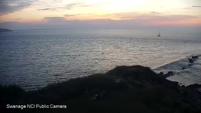 A view of the ocean at sunset, with calm waves and a sailboat in the distance. The sky is partly cloudy with soft pink and orange hues. Rocky shoreline and a small grassy area with a bench are visible in the foreground. The image is labeled "Swanage NCI Public Camera."