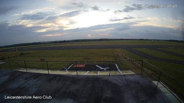 An outdoor view from a webcam at Leicester Aero Club shows an early morning sky with scattered clouds, a runway in the middle distance, and grassy fields surrounding the airfield. There is a small landing strip with a red and white marking painted on it in the foreground, edged by a metal railing. The timestamp indicates the time is 5:00 AM on July 1, 2025.