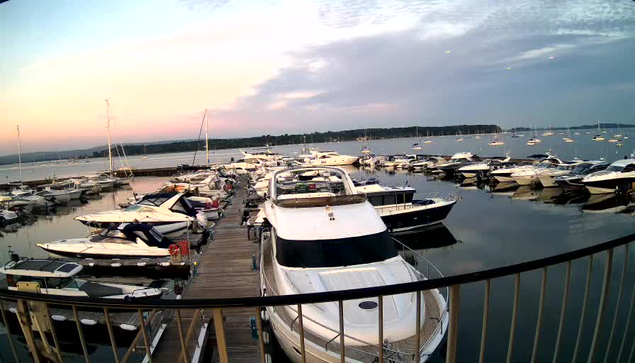 A marina filled with various boats and yachts docked along wooden piers, with calm water reflecting the boats. The sky shows a soft, colorful sunset with pink and blue hues, and a distant shoreline is visible on the horizon. A black railing curves around the foreground.