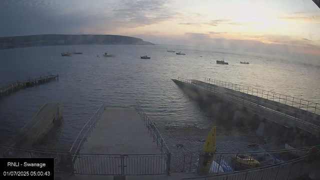 A view of the harbor at sunrise with several boats floating on calm water. There are cliffs in the distance, and the sky is partly cloudy with soft, warm light from the rising sun. In the foreground, there is a small concrete ramp and safety railings, with some equipment and a yellow caution sign nearby. The date and time are displayed in the bottom left corner as July 1, 2025, at 5:00 AM.