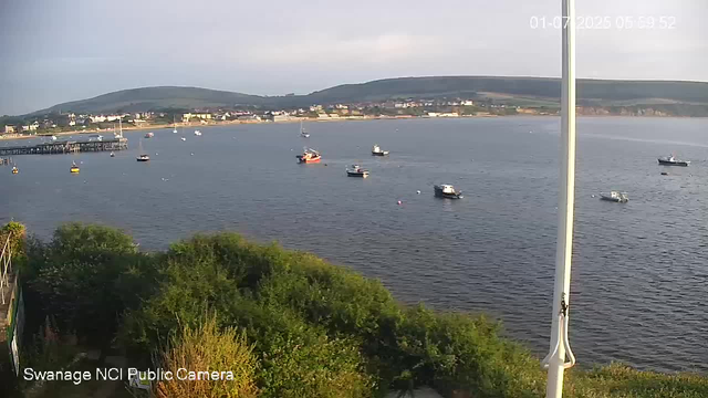 A scenic view of a harbor with several boats floating on calm water. In the foreground, there is green foliage and a white flagpole. In the background, there are hills with scattered buildings and a partly cloudy sky. The timestamp on the top right corner reads "01-07-2026 05:09:52," and the bottom left corner indicates the image is from the "Swanage NCI Public Camera."