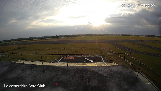 A view from a webcam at Leicesterhire Aero Club showing an airfield with a runway extending into the distance under a partly cloudy sky. The sun is low on the horizon, casting bright light and long shadows. There is a fenced grassy area and a small helipad in the foreground, with some equipment or structures nearby. The scene is calm and quiet with no visible aircraft or people.