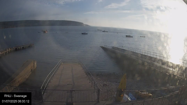 A peaceful harbor scene with a concrete slipway leading into calm water, surrounded by a metal railing. Several boats are anchored in the distance, and a large landmass with cliffs is visible across the water. The sky is partly cloudy with sunlight reflecting brightly on the water's surface. The timestamp indicates the photo was taken early in the morning.