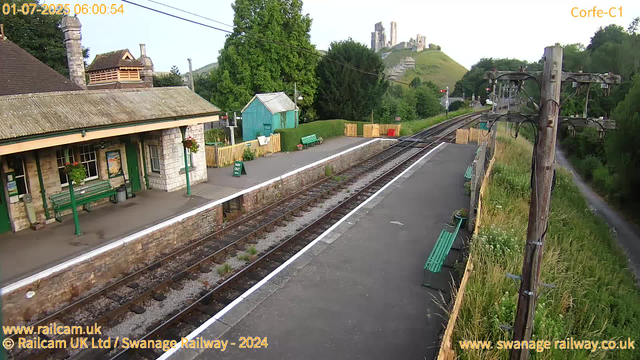 A small railway station platform with a stone building on the left side featuring green benches, flower baskets, and a "WAY OUT" sign. There are train tracks running through the center, with some grass and plants growing between the tracks. A teal-colored shed and a green bench are situated behind the platform. To the right, a wooden fence lines the edge of the platform, with several green benches and potted plants nearby. On the far right, there are tall wooden poles with electrical wires. In the background, lush green trees, a hillside, and an ancient castle ruin are visible atop the hill. The sky appears to be early morning or late afternoon. The timestamp on the image indicates it was taken on July 1, 2025, at 06:00:54.
