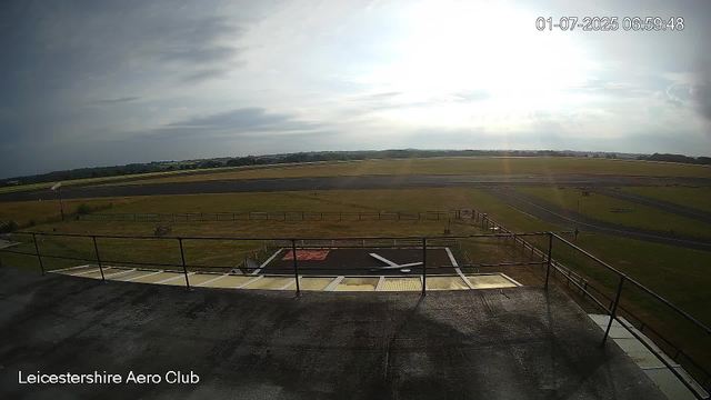 An outdoor view from a webcam at Leicesterhire Aero Club, showing a runway and grassy areas under a partly cloudy sky with bright sunlight. The timestamp reads "01-07-2025 06:59:48."