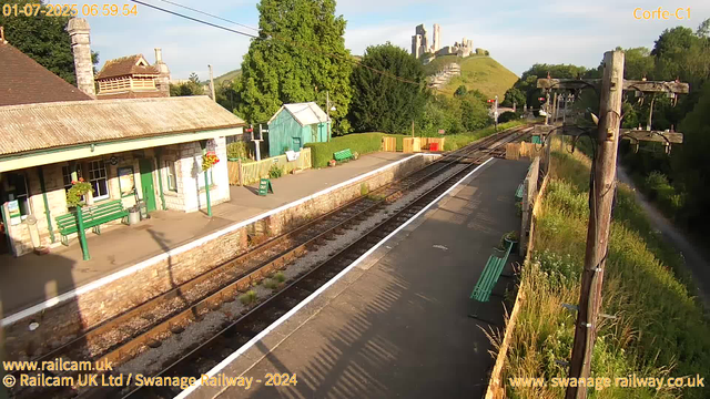 A small railway station platform with a building, green benches, and flower pots. The tracks run alongside the platform, with some railway signals and power lines overhead. In the background, there is a lush green hillside with a castle ruin on top. The scene is brightly lit with sunlight, and shadows are cast on the platform. There are some trees and bushes surrounding the area. The image has a timestamp in the upper left corner, indicating it was taken early in the morning.