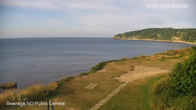 A scenic view of a coastal landscape with a calm sea stretching to the horizon. In the foreground, there is a grassy area with a small pathway made of stepping stones. To the right, dense green bushes are visible, and in the background, there is a tree-covered cliffside extending into the water. The sky is mostly clear with a few scattered clouds, and the overall scene appears peaceful with the sunlight casting gentle shadows. The timestamp in the top right corner indicates the date and time as July 1, 2025, at 6:59 AM.