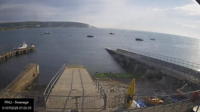 A view of a harbor from a webcam. There are several boats moored at the piers on the left side, and more boats are visible floating on the water in the distance. A breakwater extends into the water on the right side, with a sloping surface leading down to a rocky shoreline. In the background, there are cliffs along the coastline under a partly cloudy sky. The timestamp indicates it is early morning.