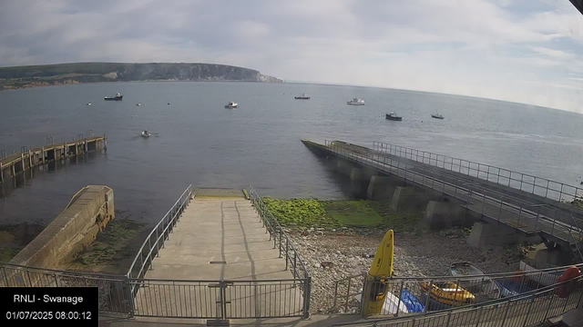 A view of a seaside scene with several boats anchored in the water. In the foreground, there is a concrete pier and a sloped ramp with railings leading into the water. To the right, a small yellow kayak and some equipment are stored near the bottom of the ramp. The sky is partly cloudy, and a distant landmass with cliffs is visible in the background across the water. A weatherproofed timestamp in the lower left corner shows the date and time.