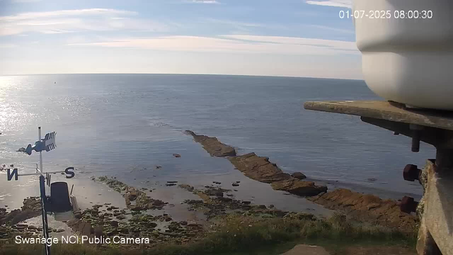 A view of a calm ocean with a rocky outcrop extending into the water. The sky is partly cloudy with sunlight reflecting off the surface of the ocean. In the foreground, there is a weather vane and an anemometer mounted on a structure. The timestamp on the image indicates the date and time as July 1, 2025, at 8:00 AM.