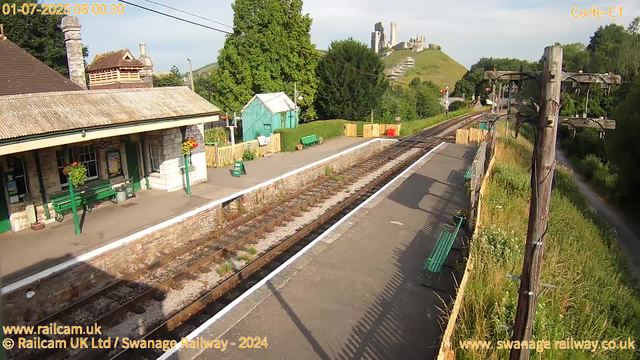 A small, rural train station with a single platform. There are green benches and hanging flower baskets on the station building, which has a sloped roof and stone walls. The platform is empty, with a "Way Out" sign and some potted plants near the building. To the right, there are railway tracks extending into the distance, with a wooden utility pole nearby that has wires attached. Behind the station, there are lush green trees and a small turquoise shed. In the background, on the hill, there are ruins of an old castle. The sky is clear with some light clouds. The timestamp in the upper left corner indicates the image was taken at 8:00 am on July 1, 2025.