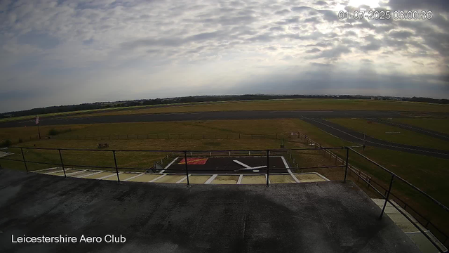 An outdoor view of an airfield taken from a webcam at Leicester Aero Club. The sky is partly cloudy with the sun partially obscured by clouds. The runway stretches into the distance with grassy areas surrounding it. In the foreground, there is a concrete platform with a railing and a small parking or staging area marked with white lines and a red symbol. A small flag is visible on the left side, and a bench is situated on the grassy area. The timestamp in the top right corner indicates the date and time: July 1, 2025, at 8:00 am.