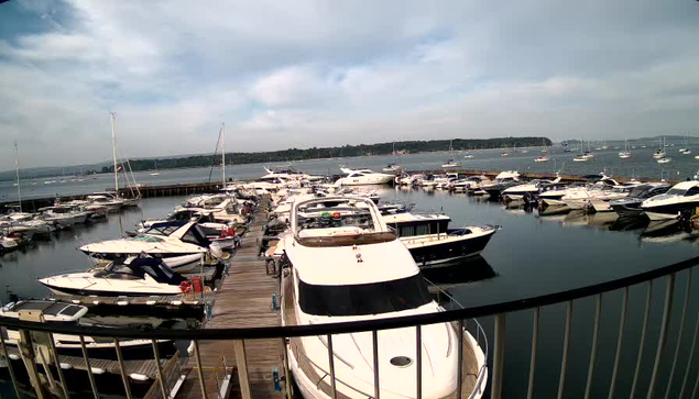A view of a marina filled with various boats and yachts docked along wooden piers, with calm water reflecting the vessels. In the background, there is a body of water extending to a distant shoreline lined with trees, under a partly cloudy sky.