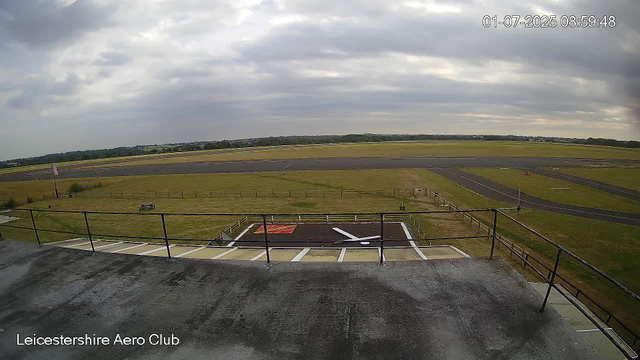 A view from a webcam at Leicestershire Aero Club shows an overcast sky with gray clouds. In the foreground, there is a building rooftop with a safety railing, and a helipad marked with a red and white circle. Beyond, there is a grassy area with a small paved pathway and a distant runway. The overall scene appears calm with no aircraft or people visible.