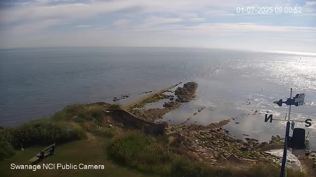 An ocean view with a rocky shoreline extending into the water, and a small grassy area with benches and bushes in the foreground. The sky is partly cloudy with sunlight reflecting off the water. There's a weather vane indicating wind direction on the right side of the image. The timestamp reads "01-07-2025 09:00:52" and the camera label in the corner says "Swanage NCI Public Camera."